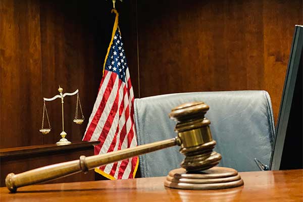 wooden-gavel-in-courtroom-on-judges-desk-with-american-flag-in-background-and-statue-of-balance-of-justice-statue-in-criminal-defense